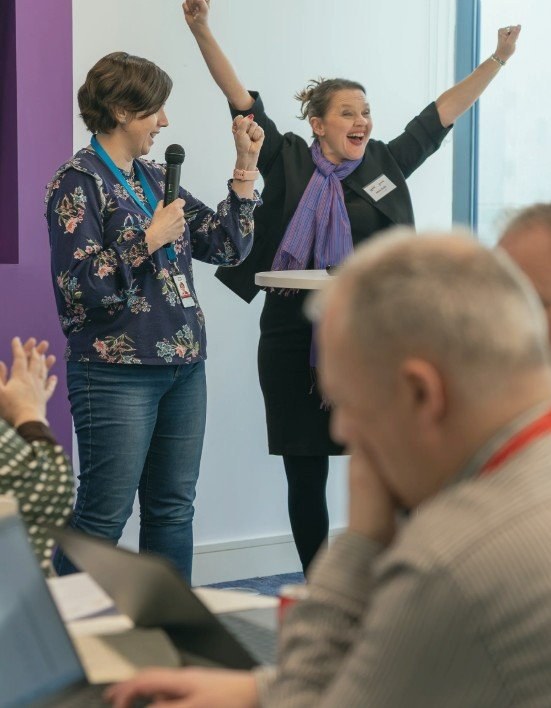 Participants at a First Prompt AI workshop, two women celebrating with raised arms, one holding a microphone, while others follow along on laptops